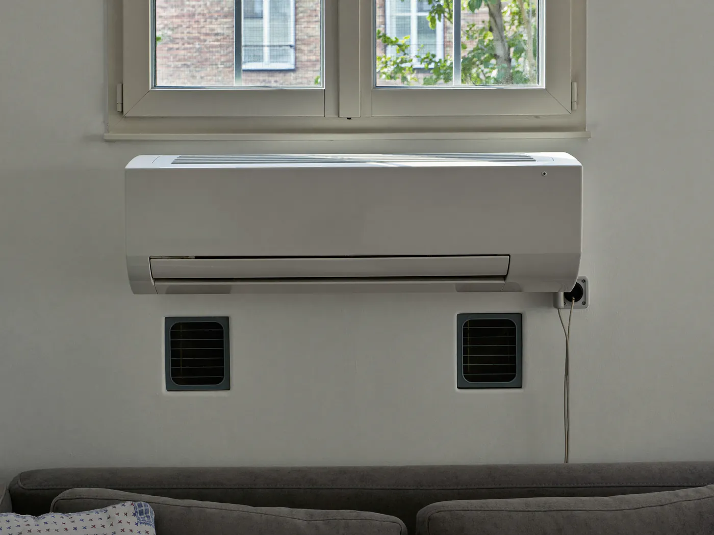 Witte airco zonder buitenunit laag tegen een binnenmuur onder het raam, met twee ronde roosters erboven