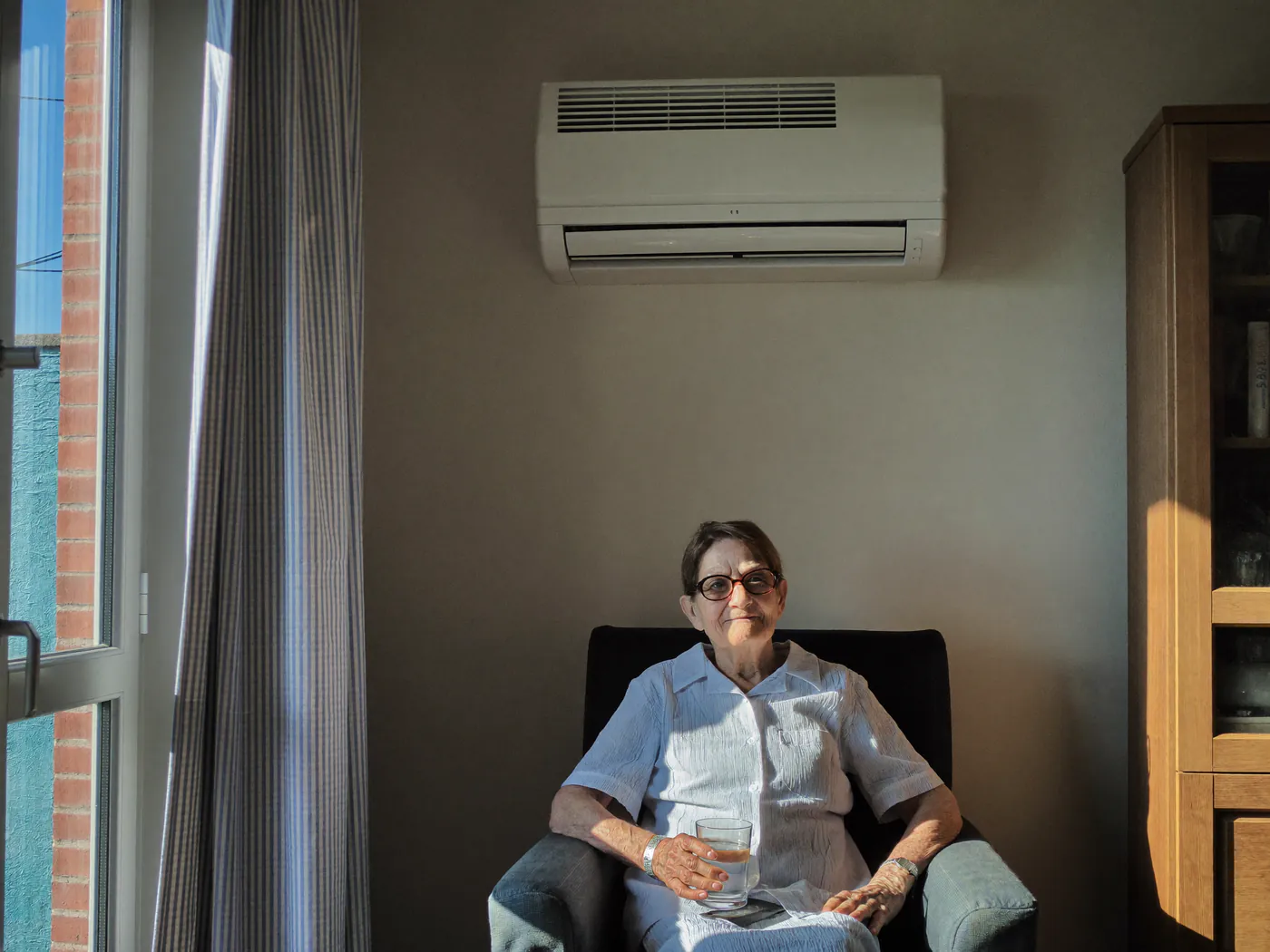 Oudere vrouw in een fauteuil met een glas water onder een binnenunit aan de wand