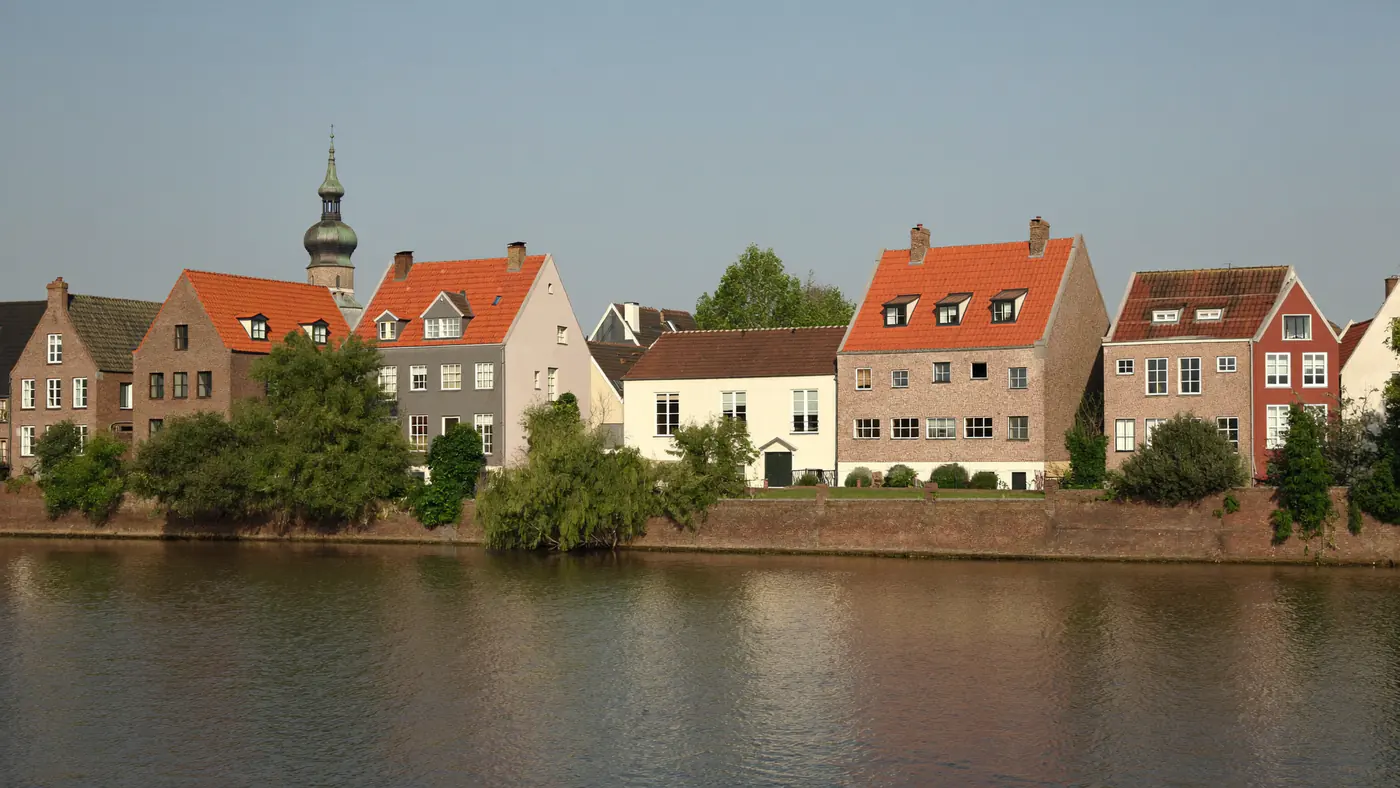 Rij oude huizen met oranje daken aan een rivier met een kerktoren erachter